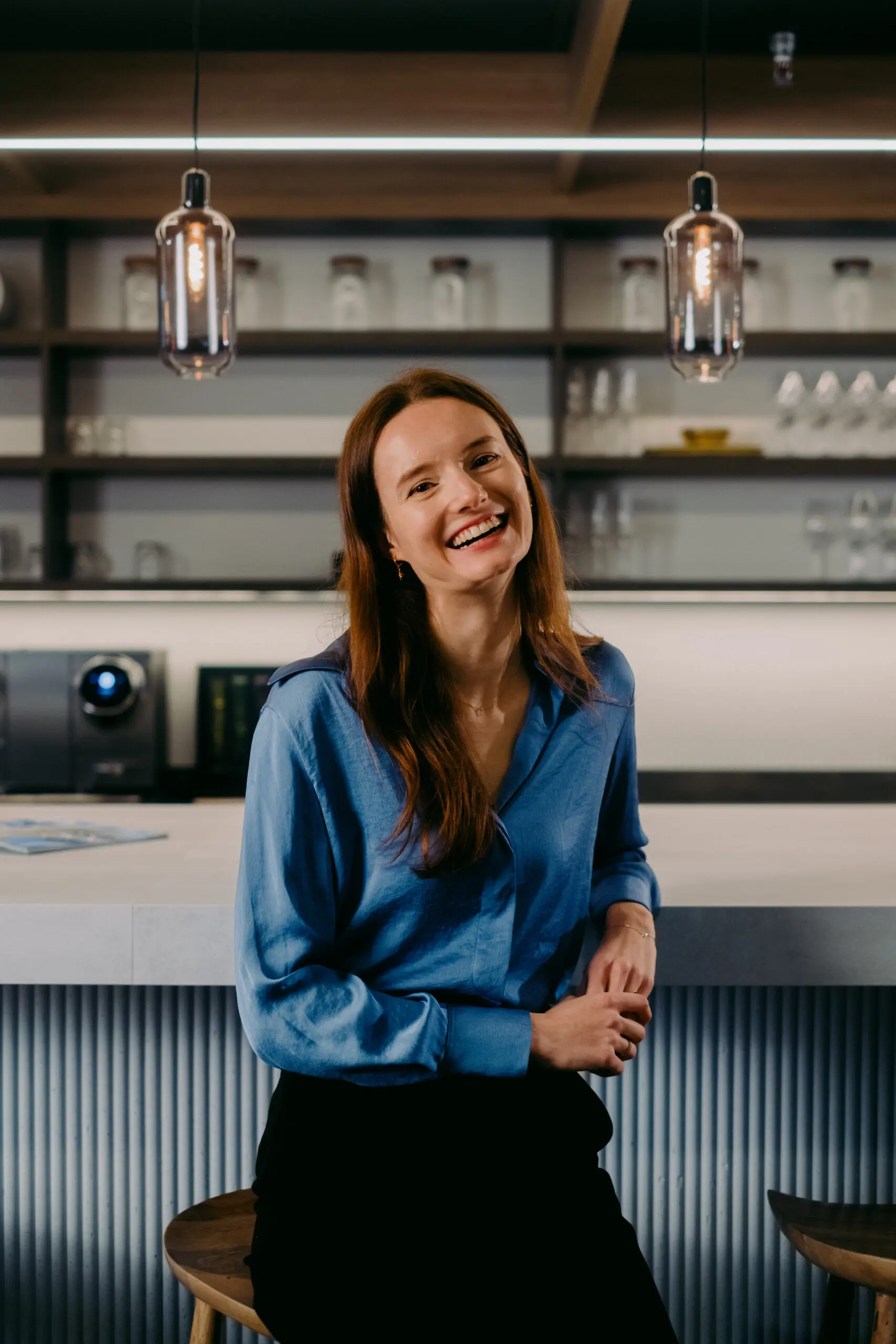 Een lachende vrouw in blauwe blouse zit aan een moderne bar met hanglampen.