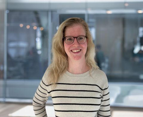 Een vrouw met bril en blond haar lacht in een moderne kantooromgeving.