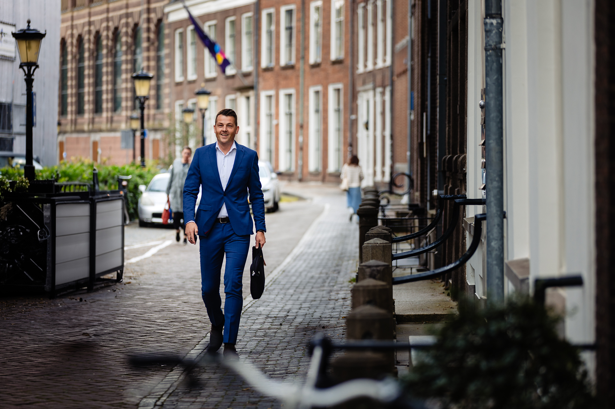 Man in blauw pak loopt met tas op straat, met historische gebouwen op de achtergrond.