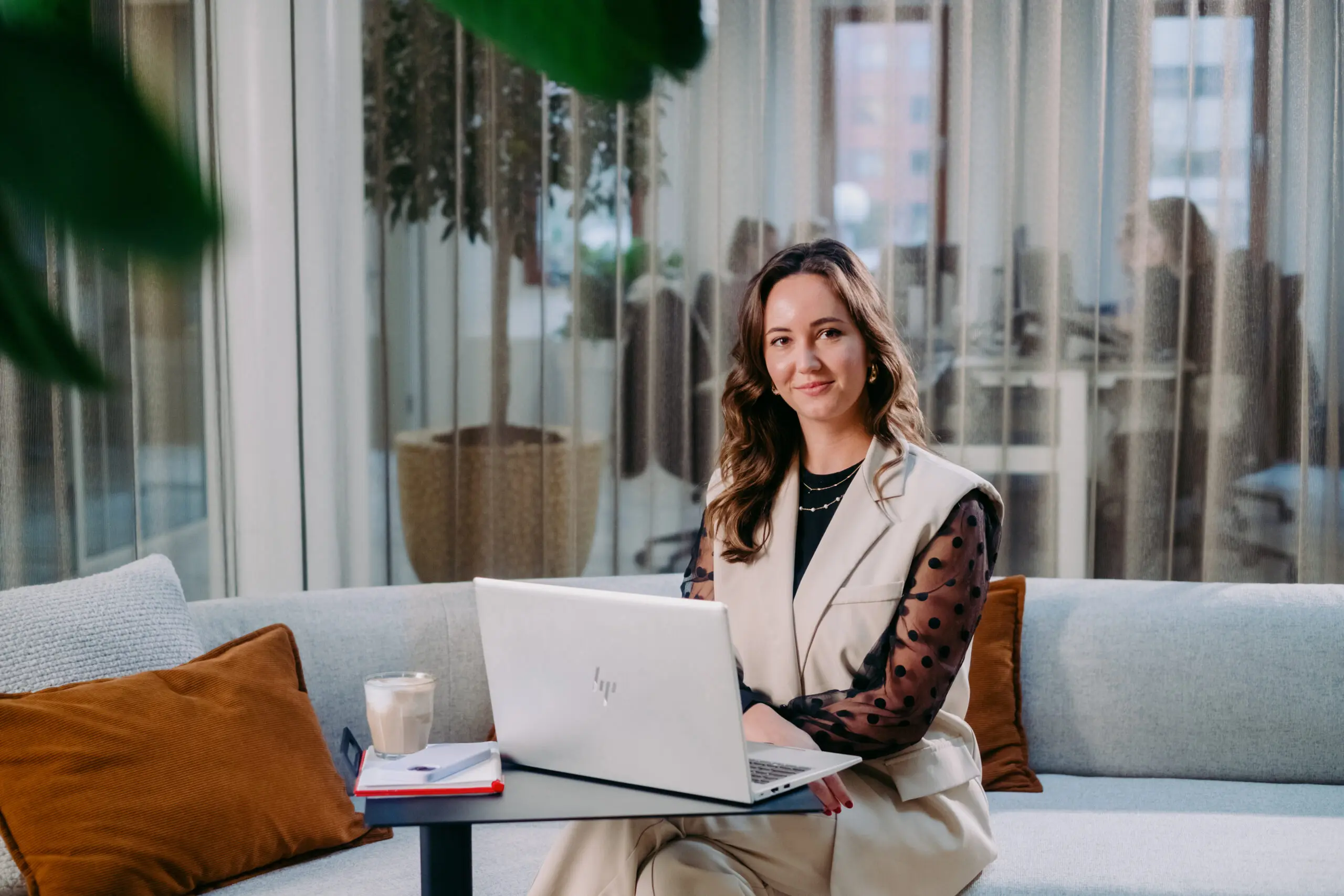 Een vrouw zit op een bank met een laptop, een kop koffie en notitieblok in een kantooromgeving.
