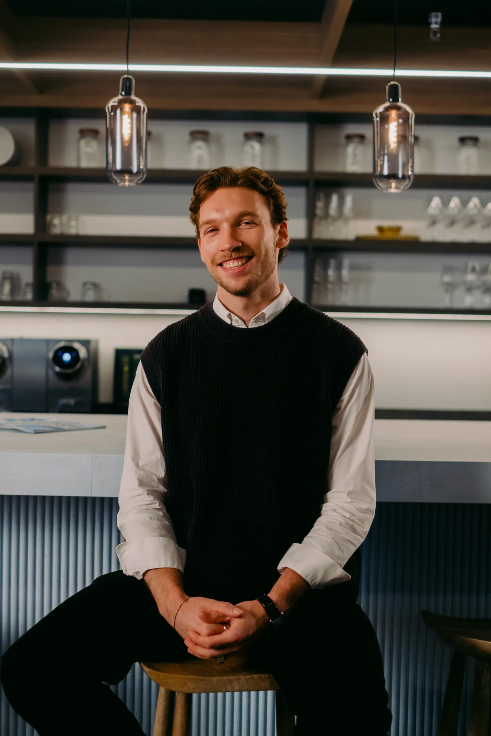 Een glimlachende man zit aan een moderne bar met hanglampen en planken vol glazen op de achtergrond.