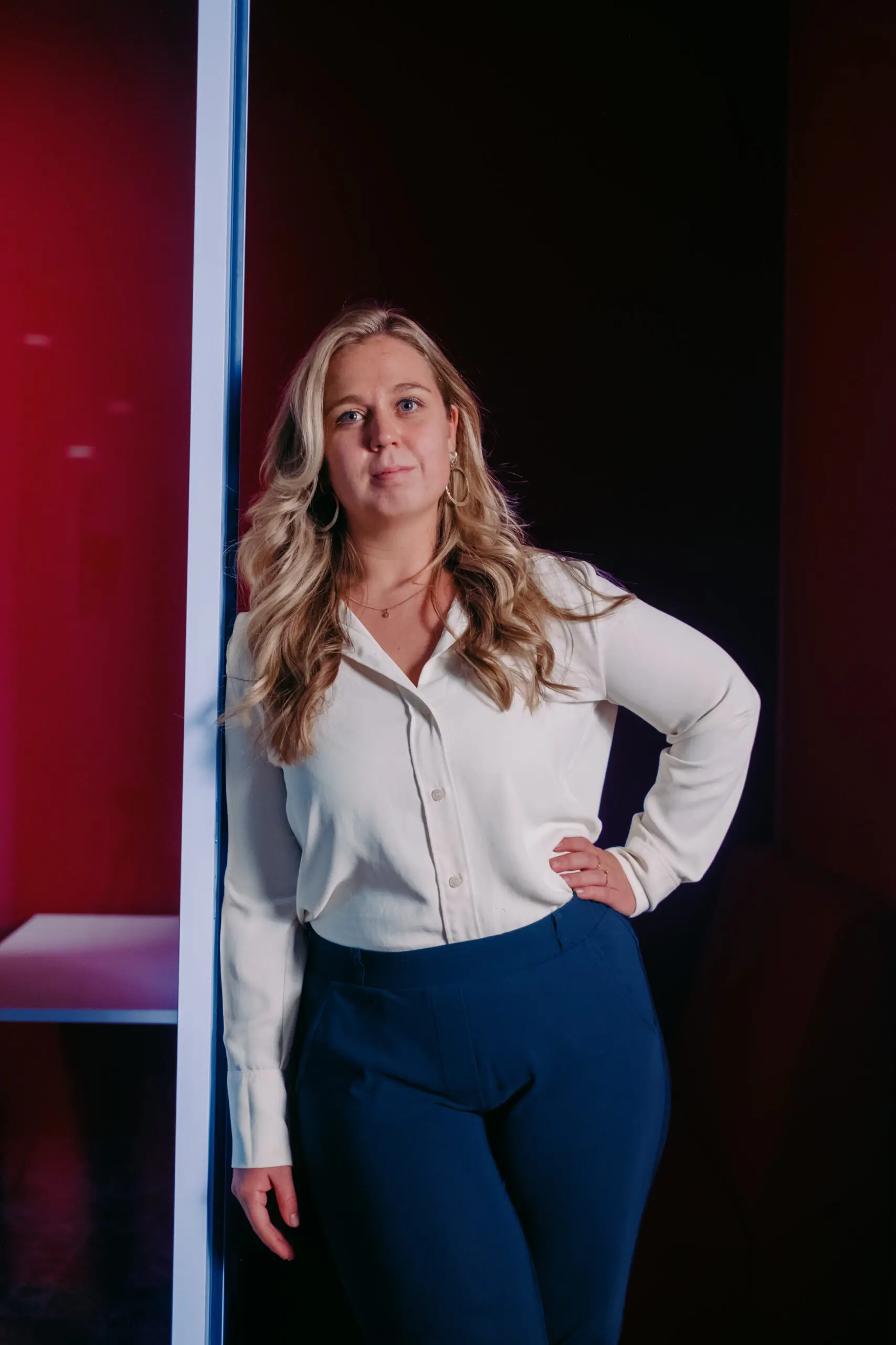 Een vrouw met blond haar, witte blouse en blauwe broek, leunt tegen een muur met een rode achtergrond.