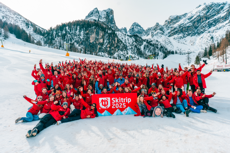 Grote groep in rode jassen met "Skitrip 2025" bord, poseren op besneeuwde berg.