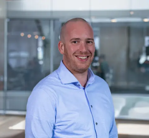 Een lachende man in een blauwe overhemd, staande voor een glazen wand in een kantooromgeving.