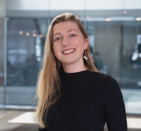 Een glimlachende vrouw met blond haar in zwart shirt, binnen in een moderne kantooromgeving.