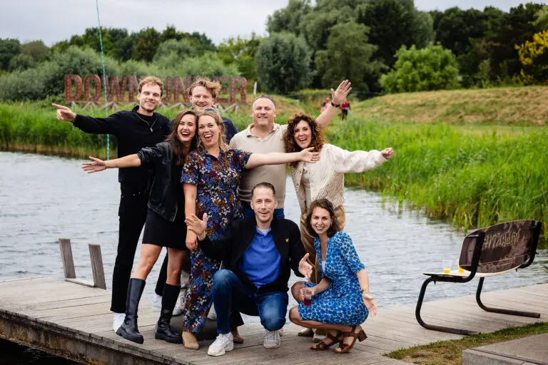 Groep mensen poseert vrolijk op een steiger bij het water, met groene natuur op de achtergrond.