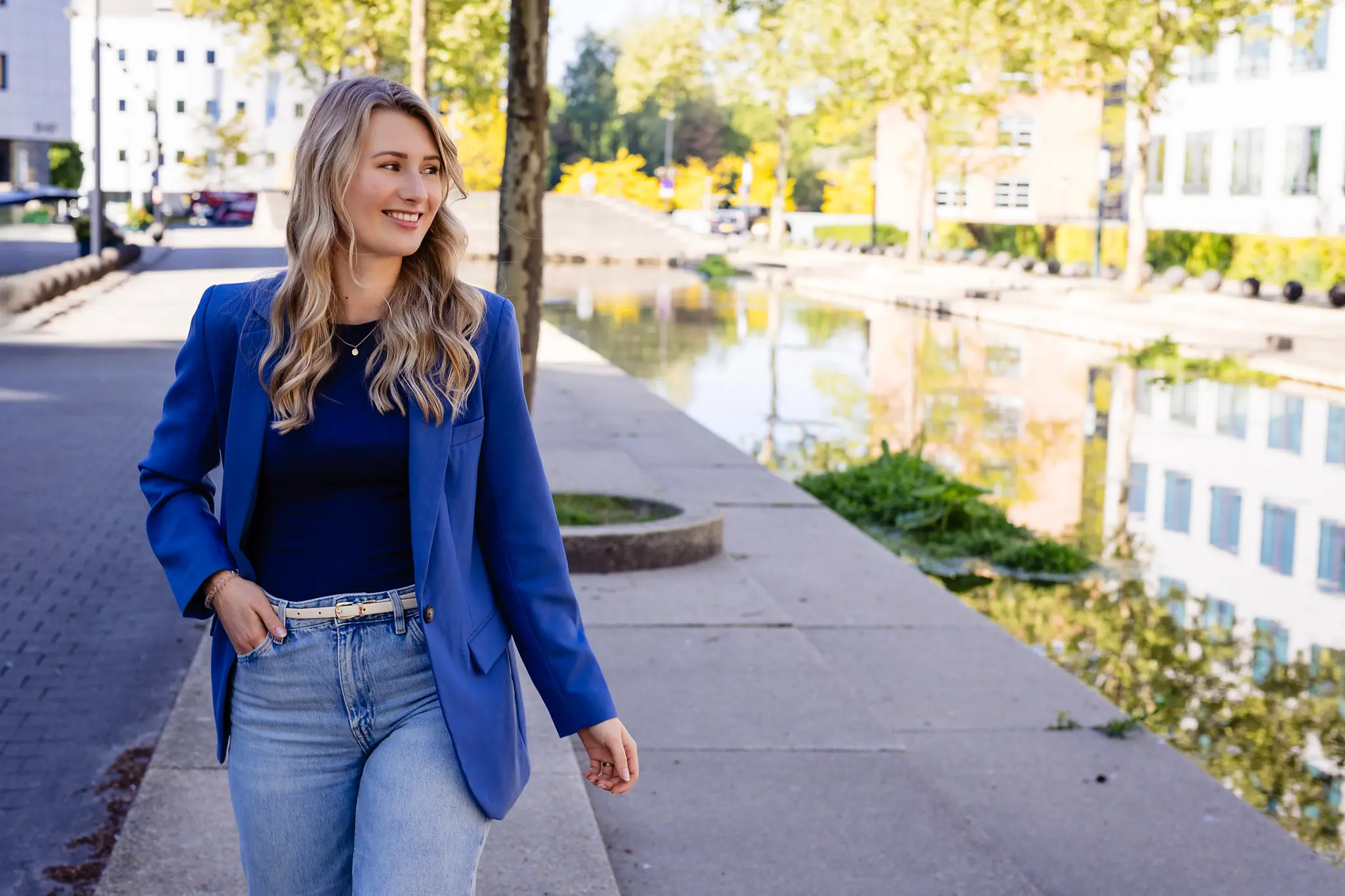 Vrouw in blauwe blazer loopt langs kanaal met bomen en gebouwen op achtergrond, zonnige dag.
