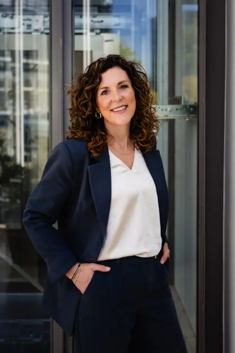 Vrouw met krullend haar in donkerblauw pak, glimlachend voor glazen gebouw.
