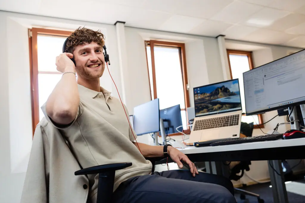 Een man met een headset lacht zittend aan een bureau met laptop en monitor.