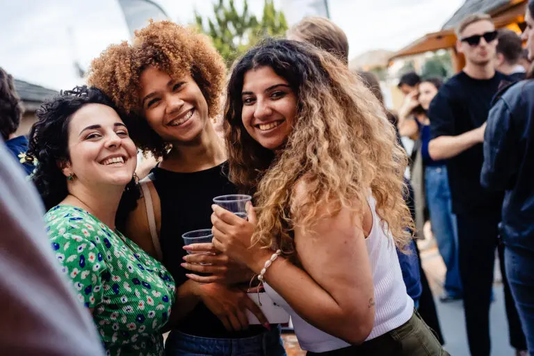 Groep lachende vrouwen met drankjes op een buitenfeest, omringd door mensen in zomerkleding.