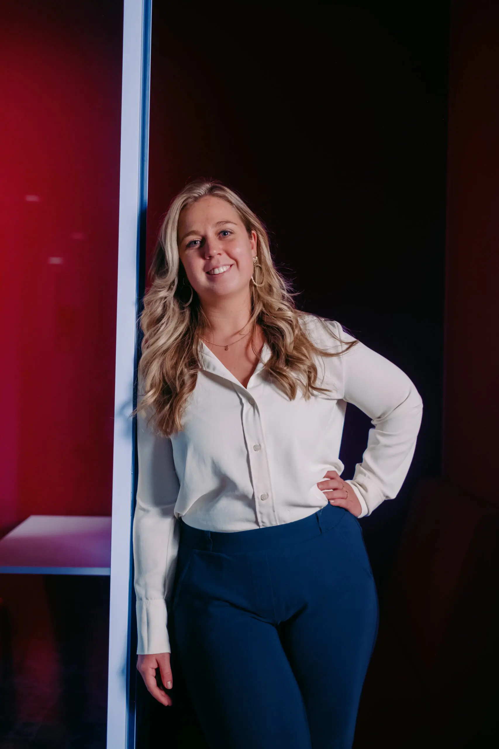 Vrouw in witte blouse en blauwe broek leunt glimlachend tegen roze muur in moderne ruimte.