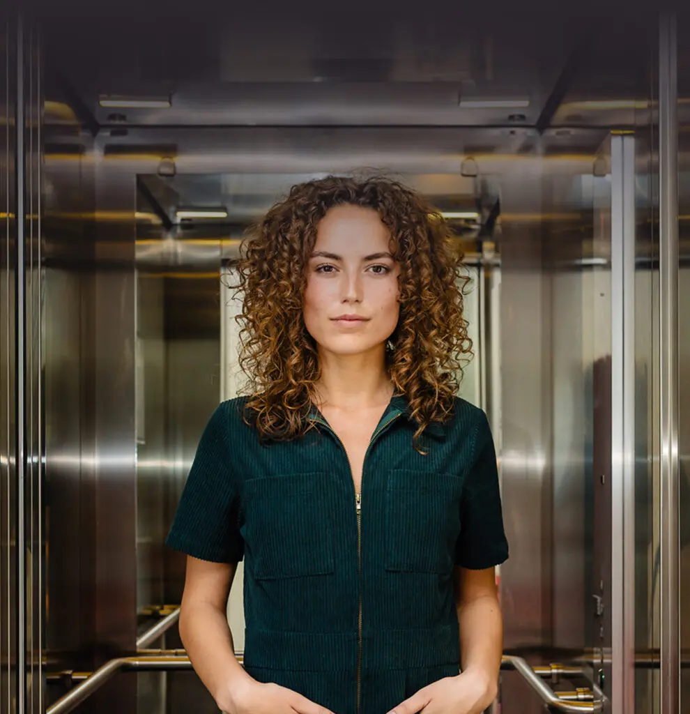 Een vrouw met krullend haar staat in een metalen lift, gekleed in een donkergroene jurk.