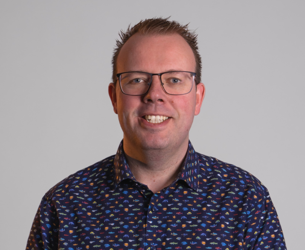 Een man met bril en gekleurd overhemd glimlacht naar de camera, tegen een neutrale achtergrond.