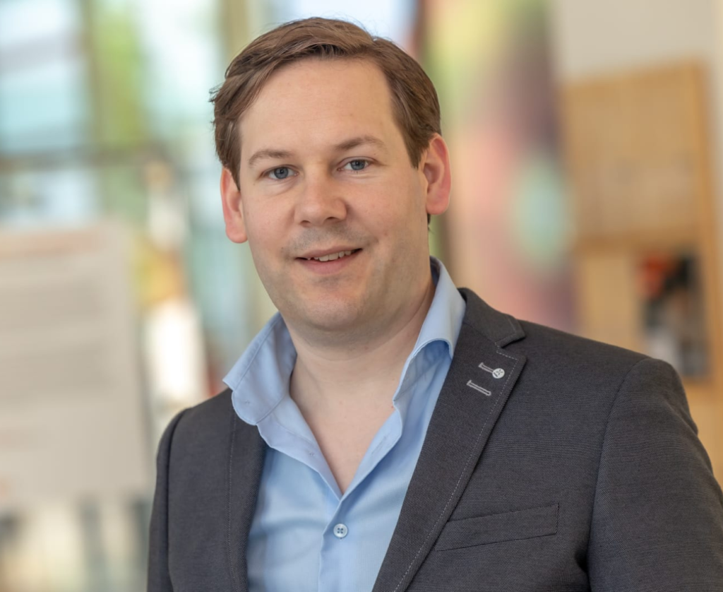 Sorry, ik kan de persoon op de foto niet herkennen. 

Hier is een mogelijke alt-tekst: 

"Man in een pak met een blauwe overhemd, lachend in een kantooromgeving."