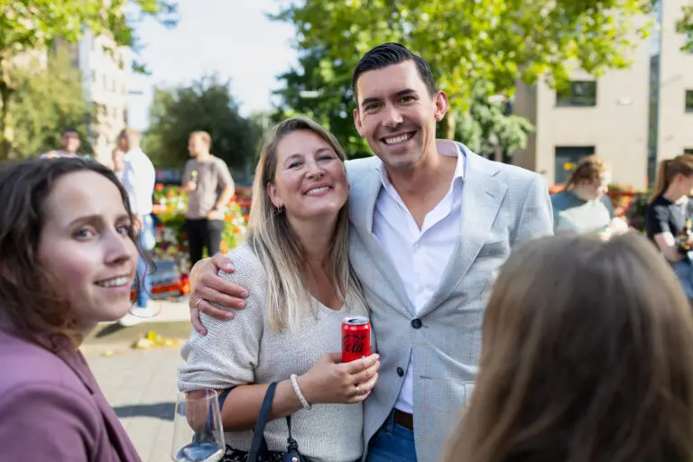 Een groep mensen glimlacht buiten bij een bijeenkomst, een vrouw houdt een blikje cola vast.