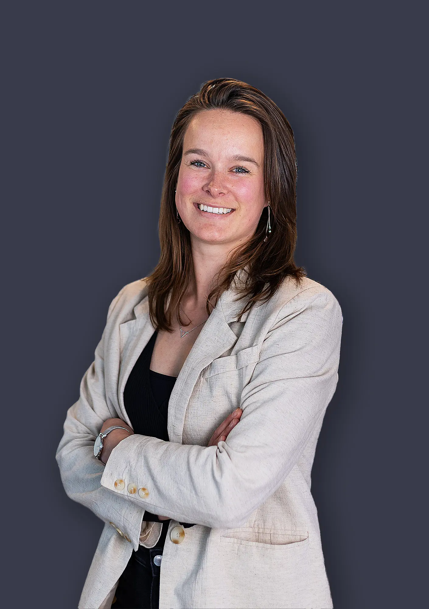 Een vrouw met lang bruin haar en beige colbert, glimlachend met gekruiste armen voor een grijze achtergrond.
