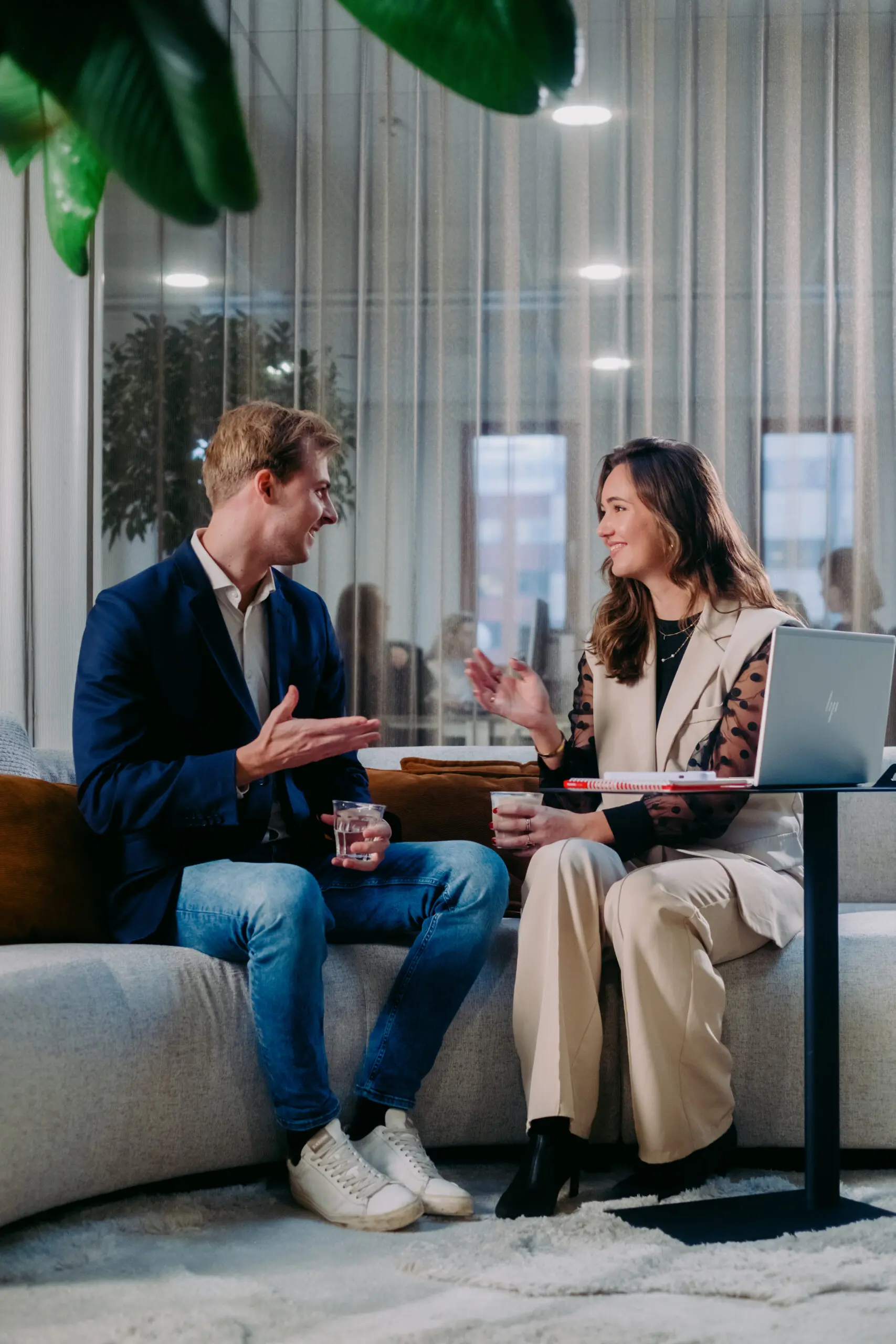 Twee mensen praten en lachen op een bank in een kantooromgeving, met laptop en notitieboekje.