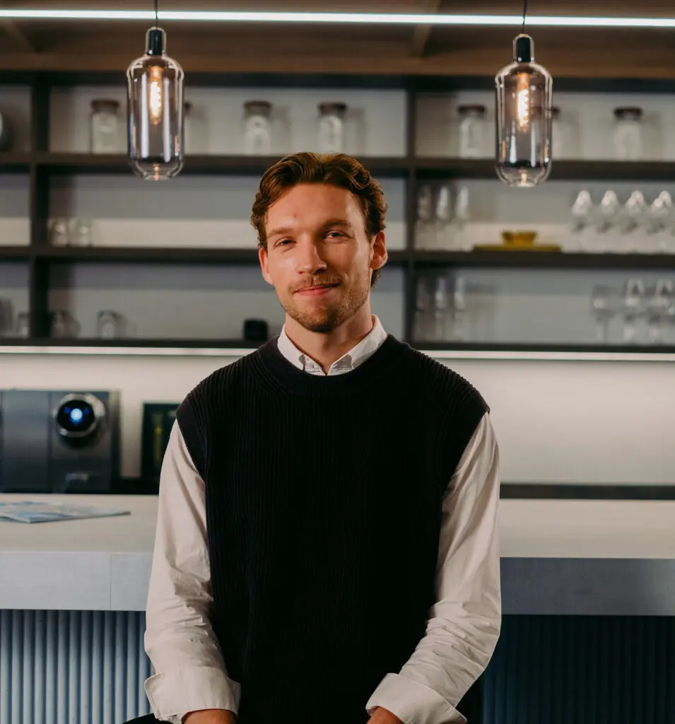 Een man poseert in een moderne keuken met hanglampen en glazen in de achtergrond.