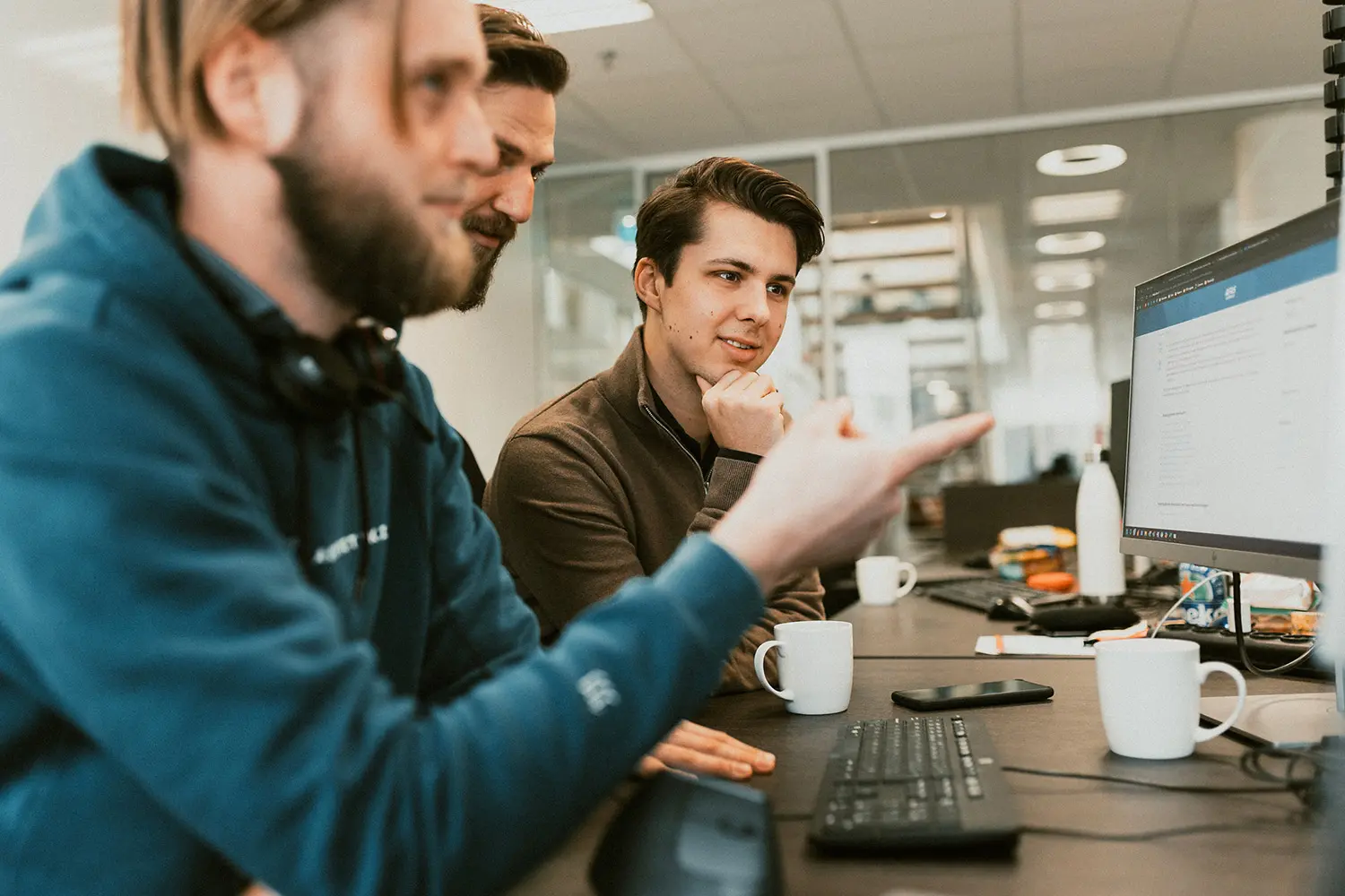 Drie mannen overleggen bij een computer in een kantooromgeving, met koffie en toetsenbord.