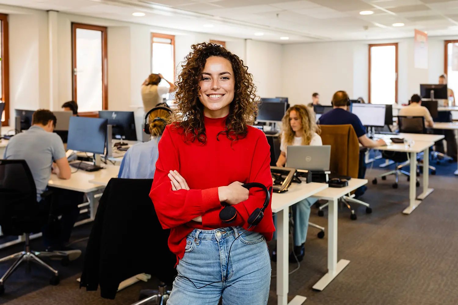 Jonge vrouw in rode trui staat glimlachend in een kantoor met werkende collega’s op de achtergrond.
