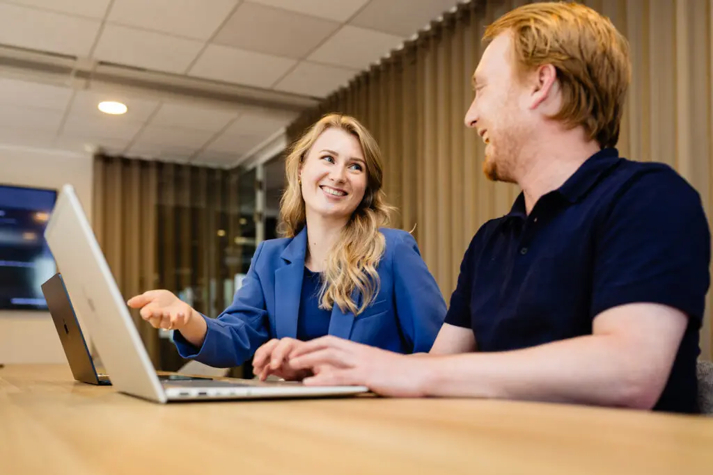 Twee mensen glimlachen en praten terwijl ze werken op laptops in een kantoorruimte.