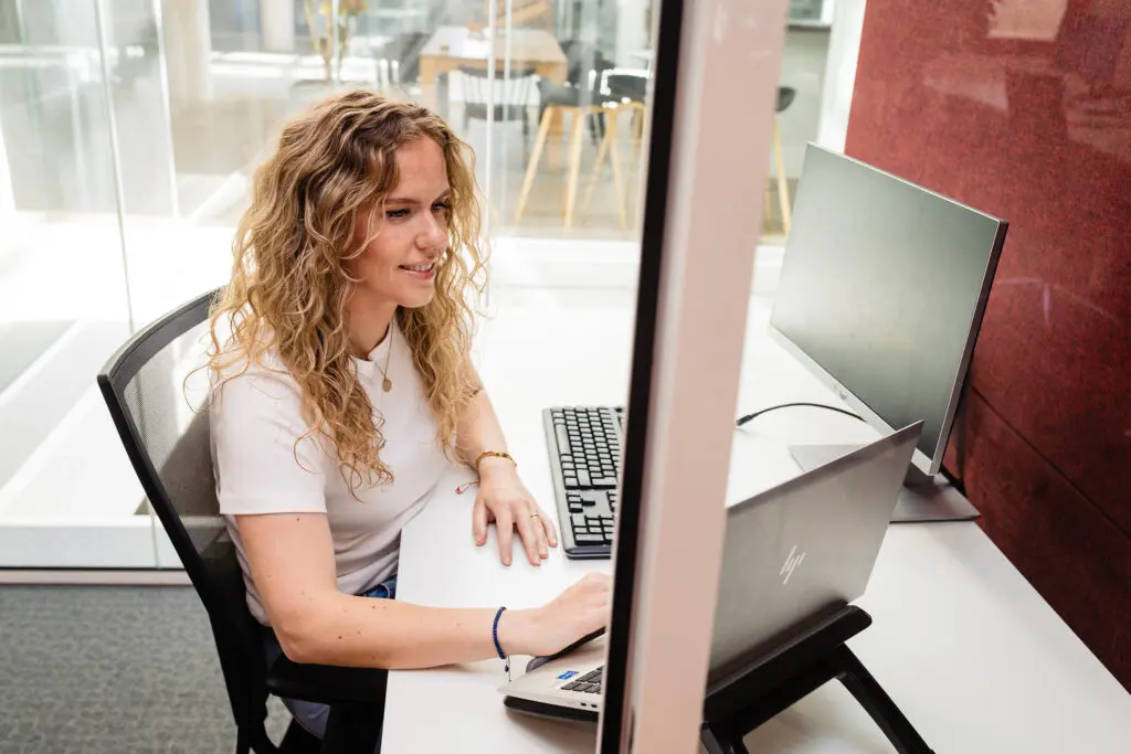Vrouw werkt glimlachend op laptop en toetsenbord in modern kantoor met glazen wand.