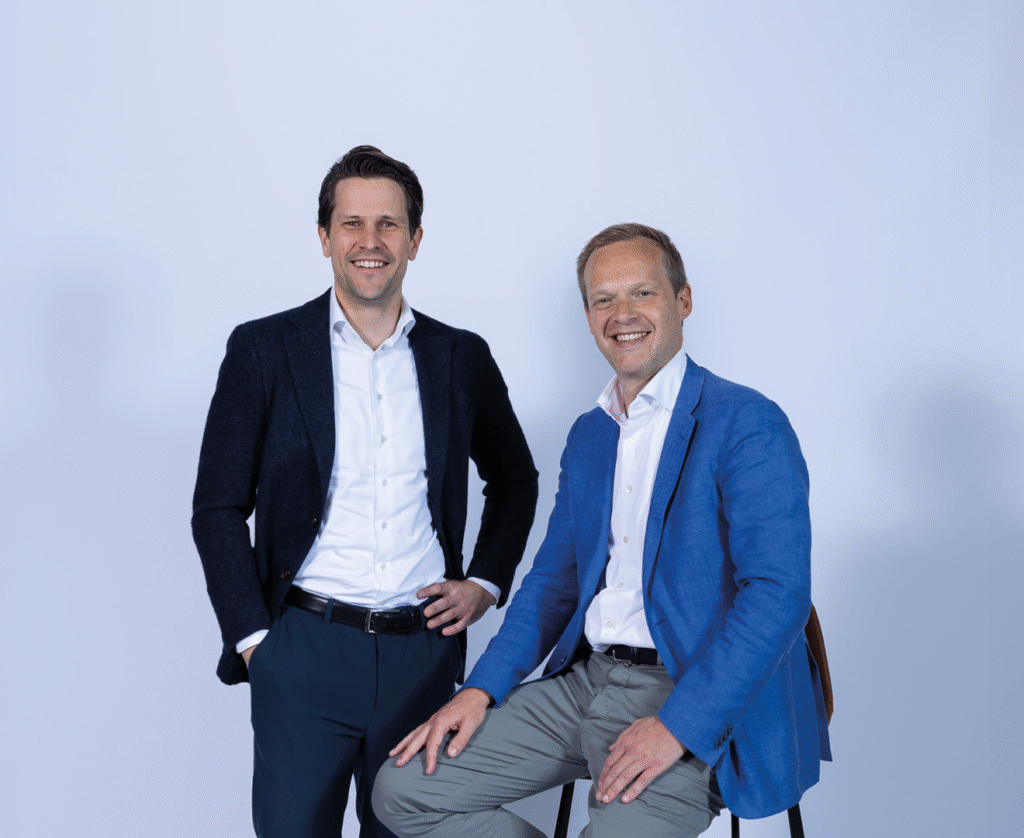 Twee mannen in pakken poseren tegen een witte achtergrond, één zittend, de ander staand.