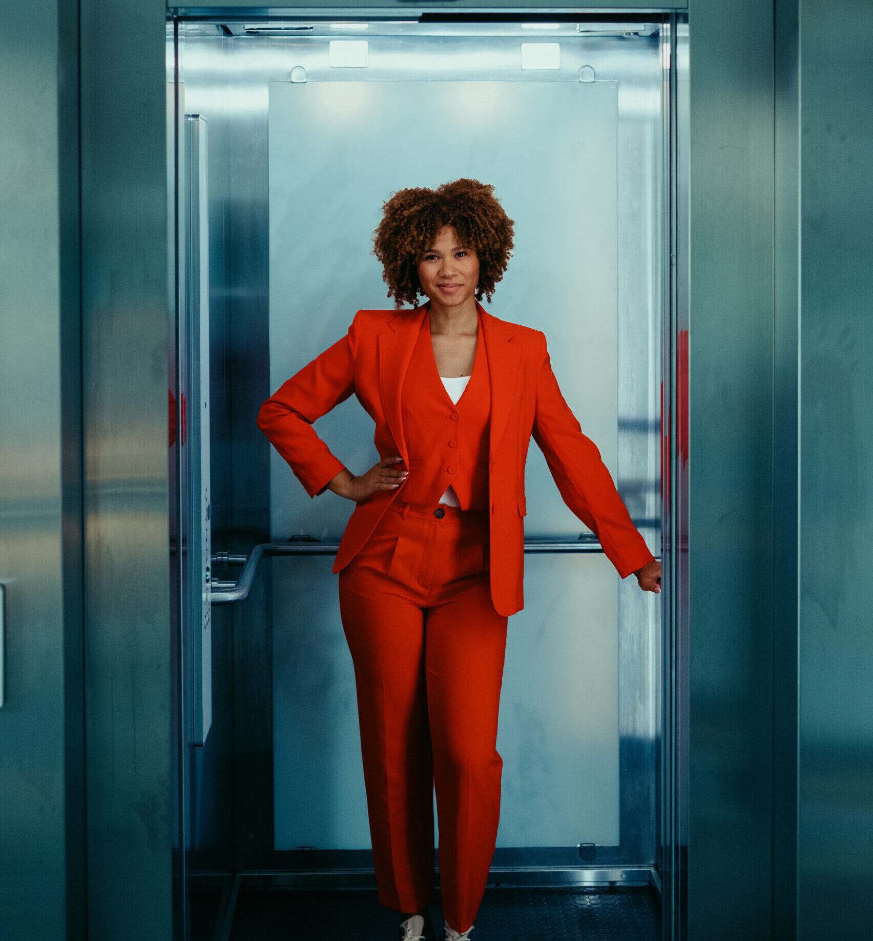 Vrouw in een rood pak staat in een metalen lift met een ontspannen houding.