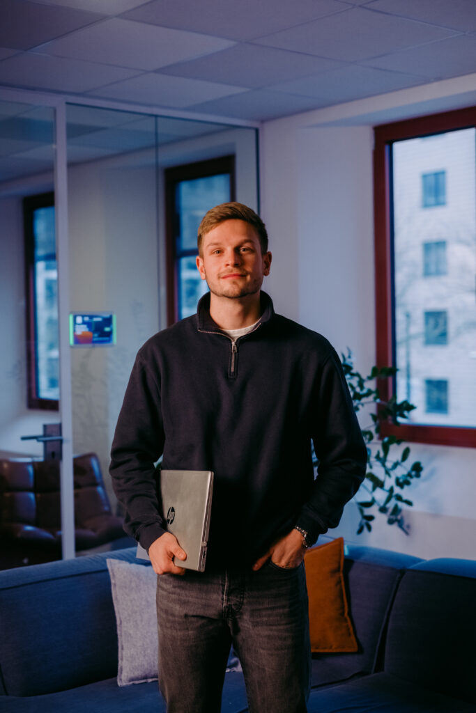 Een man in een kantoor met een laptop in zijn hand, staand voor een blauwe bank.