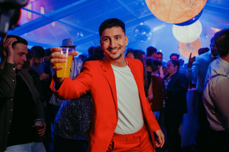 Feest met man in rood pak die een glas sinaasappelsap heft, omringd door dansende mensen.