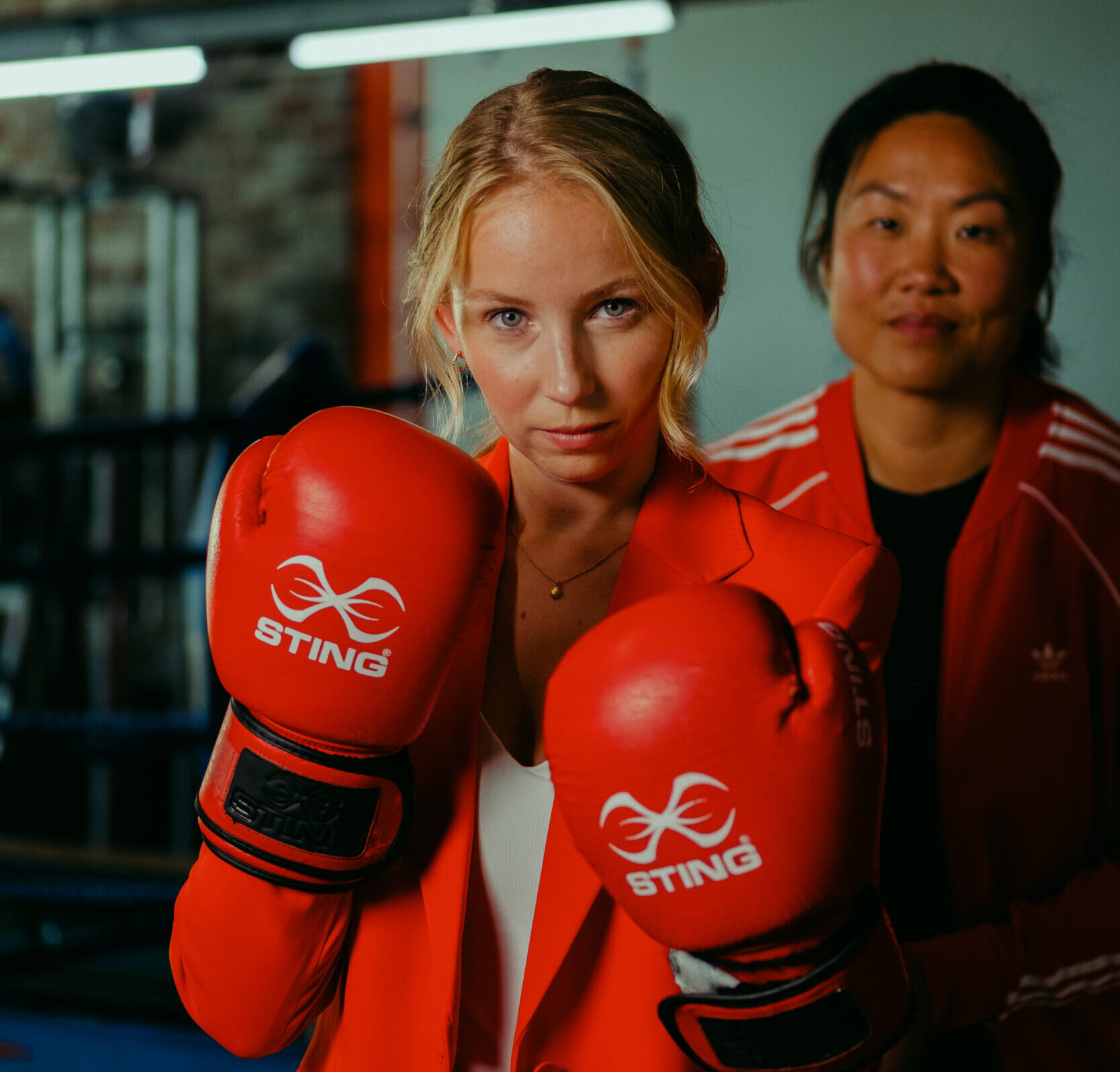 Een vrouw in een rode blazer en bokshandschoenen, met een coach op de achtergrond in een gymzaal.