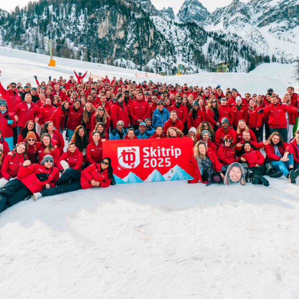 Grote groep mensen in rode jassen poseren op een besneeuwde berg met bord "Skitrip 2025".