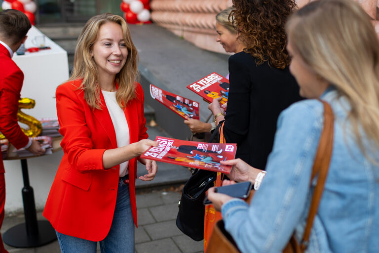 Vrouw in rode blazer deelt brochures uit aan voorbijgangers bij evenement.