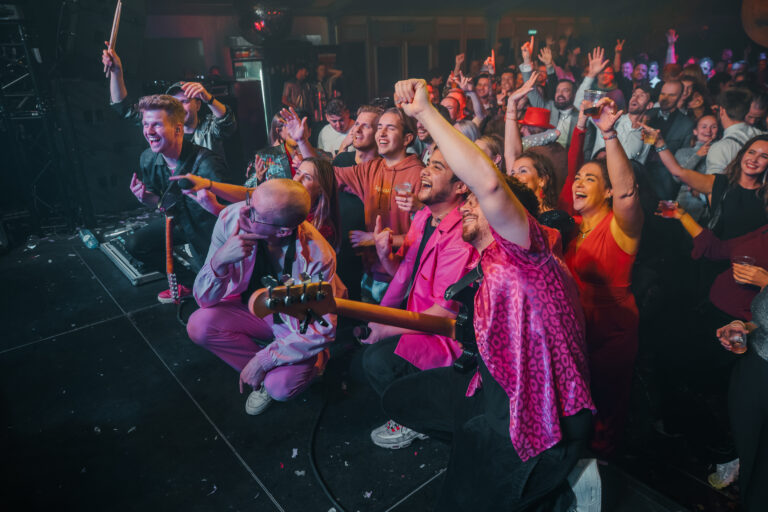 Een vrolijke menigte juicht tijdens een concert, met muzikanten in kleurrijke outfits op het podium.