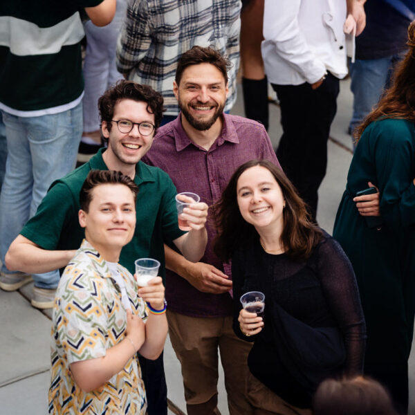Vier mensen lachen naar de camera op een druk feest, met drinken in kunststof bekers.