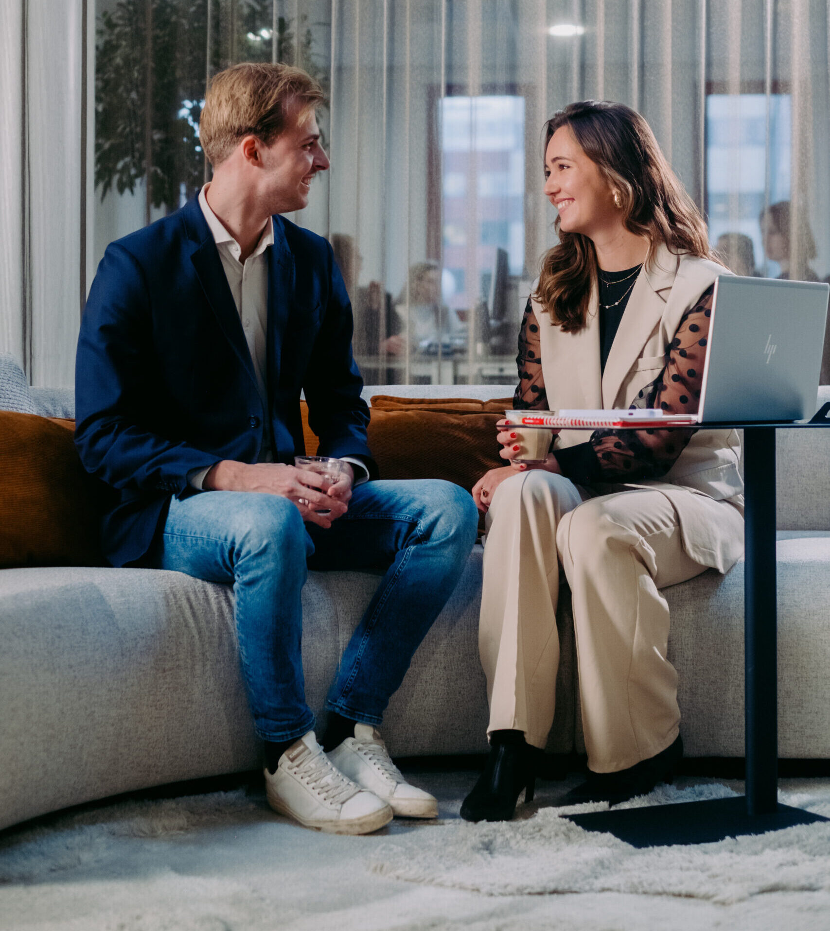 Een man en vrouw in gesprek op een bank, met laptop en drankje in een moderne kantoorruimte.