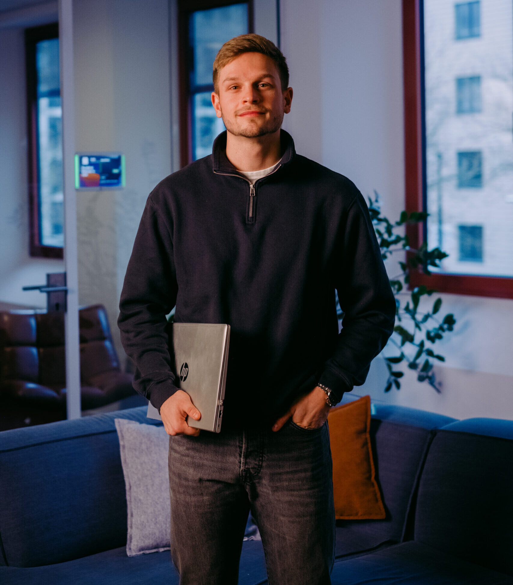 Een man in een kantooromgeving met een laptop in de hand, voor een bank met kussens.