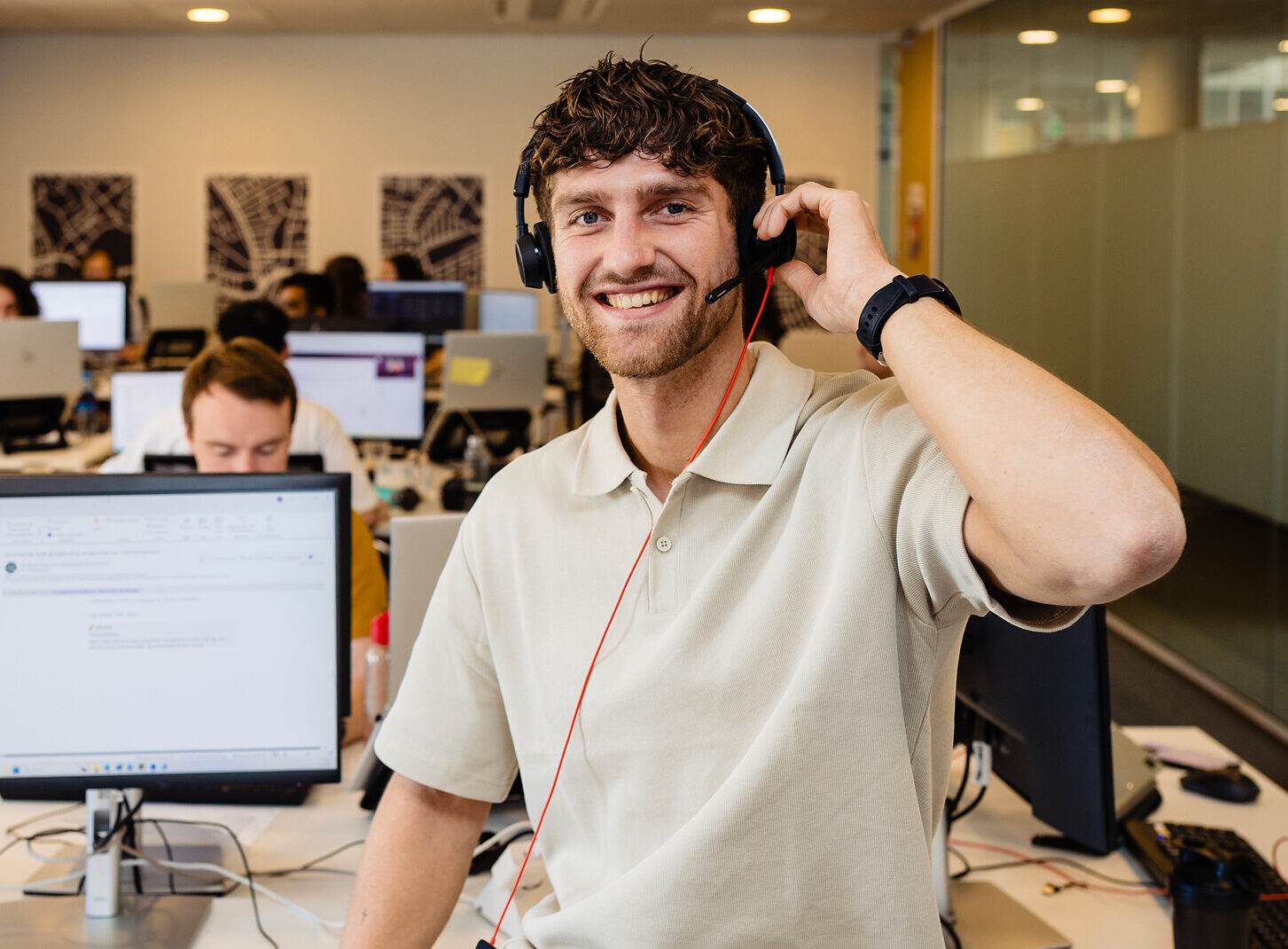 Een lachende man met headset zit op bureau in open kantoorruimte met computers.