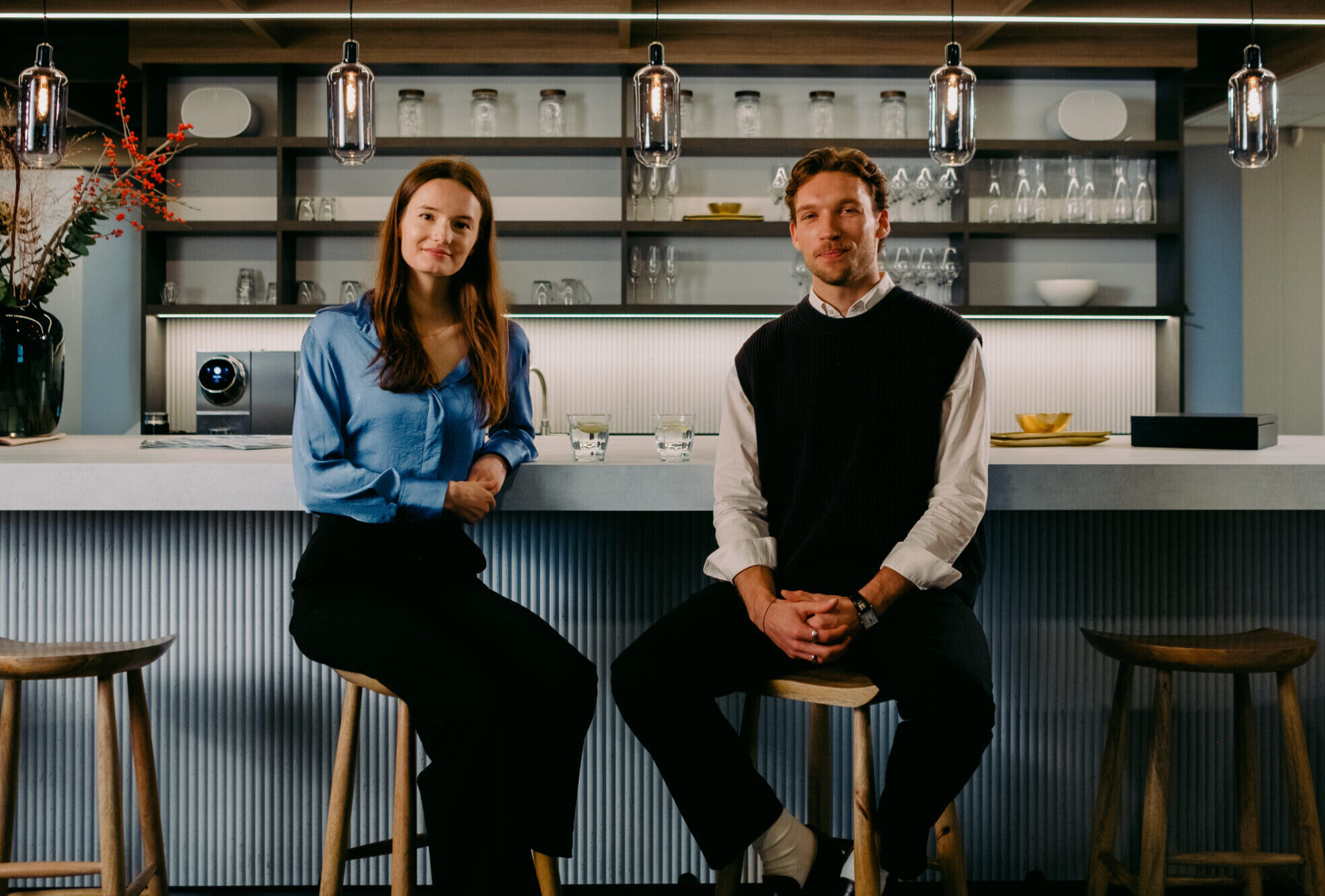 Twee mensen zitten aan een moderne bar, met glazen en hanglampen erboven.