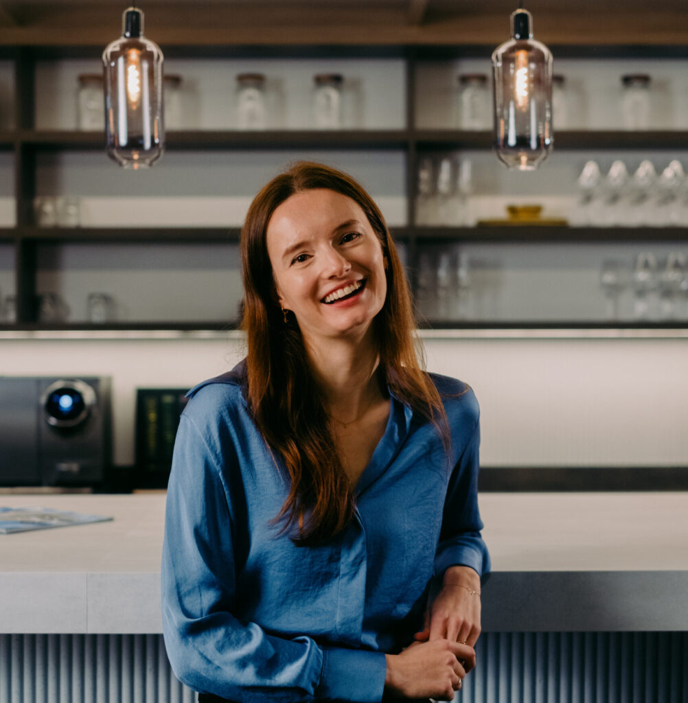 Een vrouw in een blauwe blouse lacht aan een moderne bar, met lampen en glazen op de achtergrond.